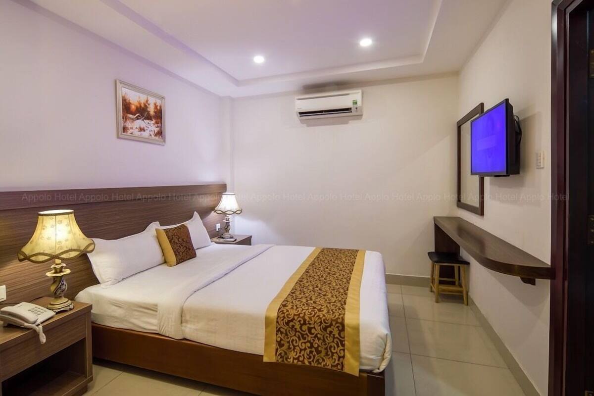 A hotel room with a bed, bedside table with a lamp, an air conditioner, a TV mounted on the wall, and a small stool.
