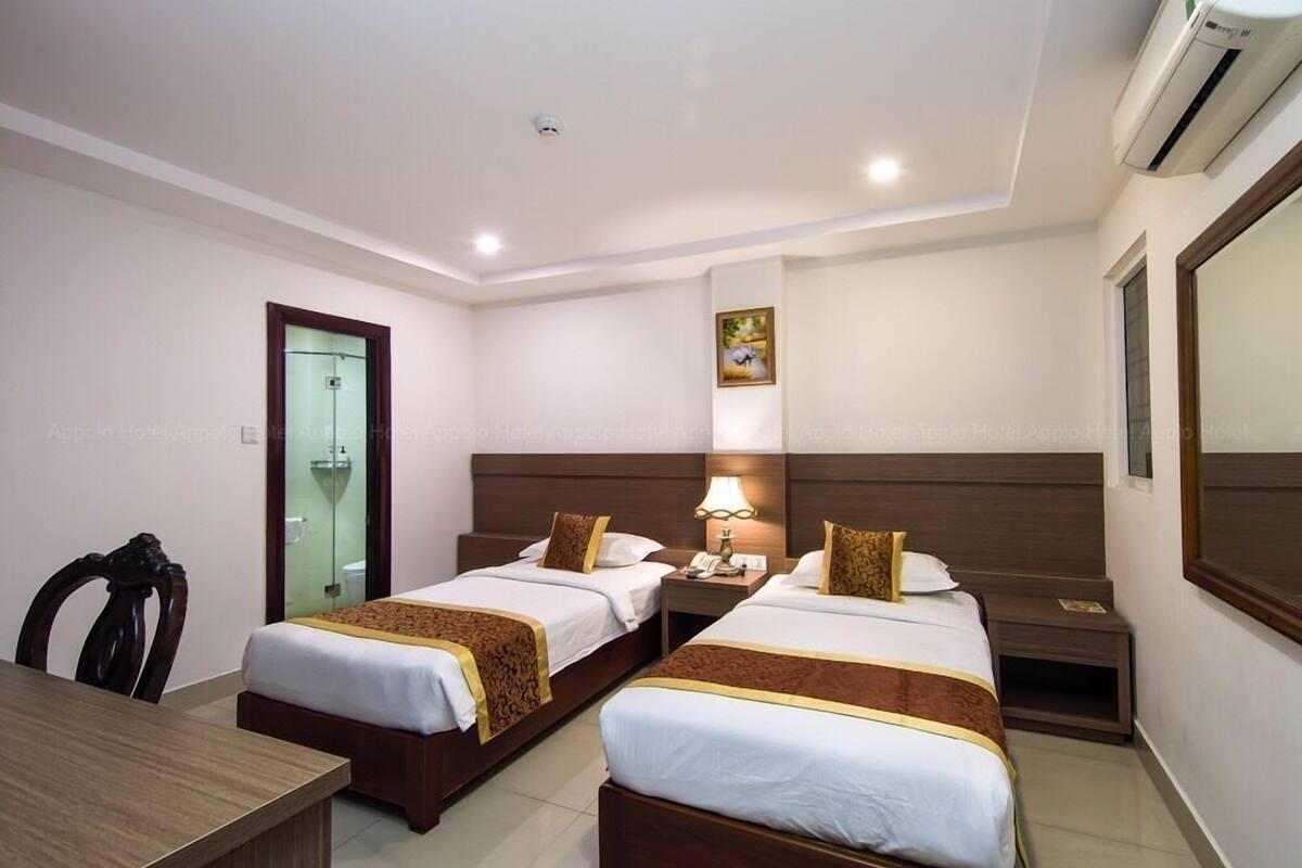 A hotel room with two single beds, a desk, a chair, a bedside table with a lamp, and a painting on the wall.