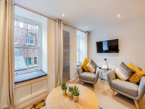 A modern living room with a large window, two armchairs, a round coffee table with plants, a TV, and a radiator. - Town Square House - UK45610 (Alnwick)