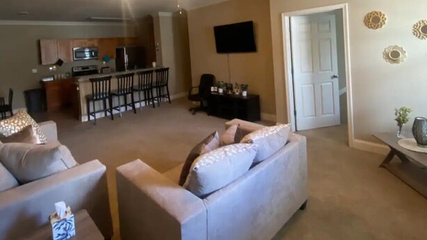 Ein Wohnzimmer mit einer Couch, einem Couchtisch und einem Fernseher. Im Hintergrund ist eine Küchenzeile mit einem Esstisch und Stühlen zu sehen.