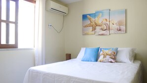 A neatly made bed with white and blue bedding, a wall-mounted air conditioner, and a window with curtains. - Casa Itacaré - Santa Onda (Itacaré)