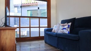 A room with a wooden desk, a blue sofa with pillows, and a large window with a view of a building and street. - Casa Itacaré - Santa Onda (Itacaré)