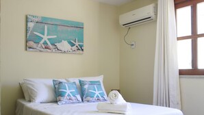 A neatly made bed with white bedding and starfish-themed pillows, an air conditioning unit on the wall, and a window with curtains. - Casa Itacaré - Santa Onda (Itacaré)