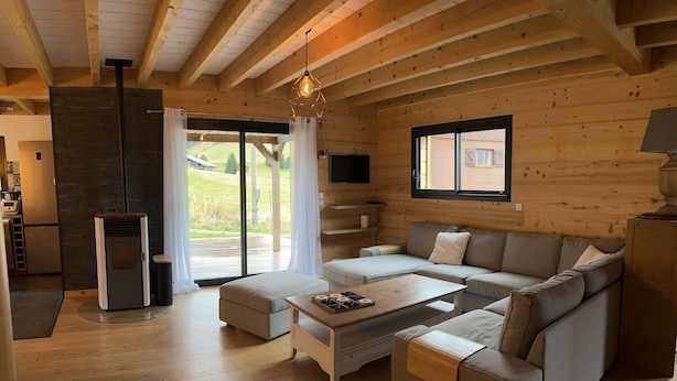 L’intérieur d’un chalet moderne en bois, avec un plancher en bois, un canapé d’angle, une table basse, une cheminée et une grande fenêtre donnant sur le paysage extérieur.
