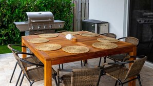 A wooden dining table with woven placemats, surrounded by wicker chairs, set outdoors with a grill and a chimney in the background. - BaeCation Dania! Near Fort Lauderdale Airport, beach & casino!
pool & jacuzzi! (Dania Beach)
