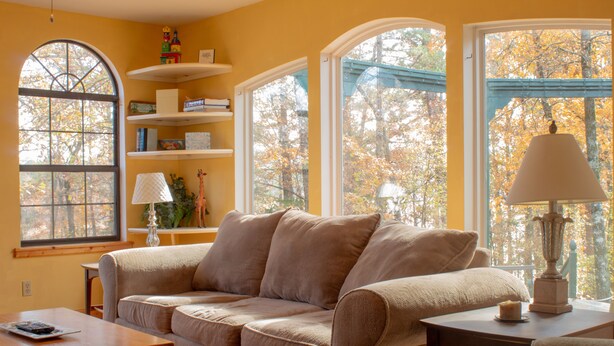 Un salón acogedor con un sofá color beige, una mesa de centro de madera y una lámpara sobre una mesita auxiliar. Hay estanterías con libros y objetos decorativos. Amplias ventanas ofrecen vistas a árboles y un cielo azul.