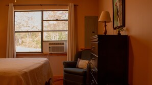 Un dormitorio con cama, una silla, cómoda, lámpara y ventana con cortinas.
