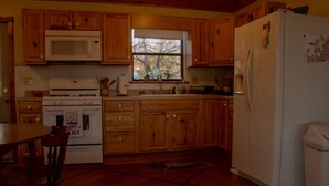 Una cocina con armarios de madera, un refrigerador blanco, un microondas, un horno y una ventana con vista a árboles.