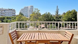 Une terrasse extérieure avec une table et des chaises en bois, une balustrade blanche et une vue sur des bâtiments et des arbres en arrière-plan.