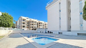 Un espace piscine avec une terrasse carrelée, un bâtiment doté de nombreux balcons et un ciel d’un bleu éclatant.