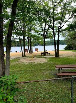 Un parque junto al lago con mesas de picnic y bancos, rodeado de árboles y una zona de playa de arena.