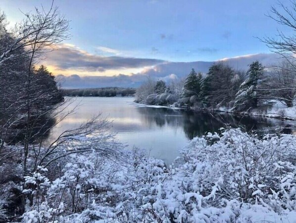 A serene winter landscape with a calm river, snow-covered trees, and a clear sky.