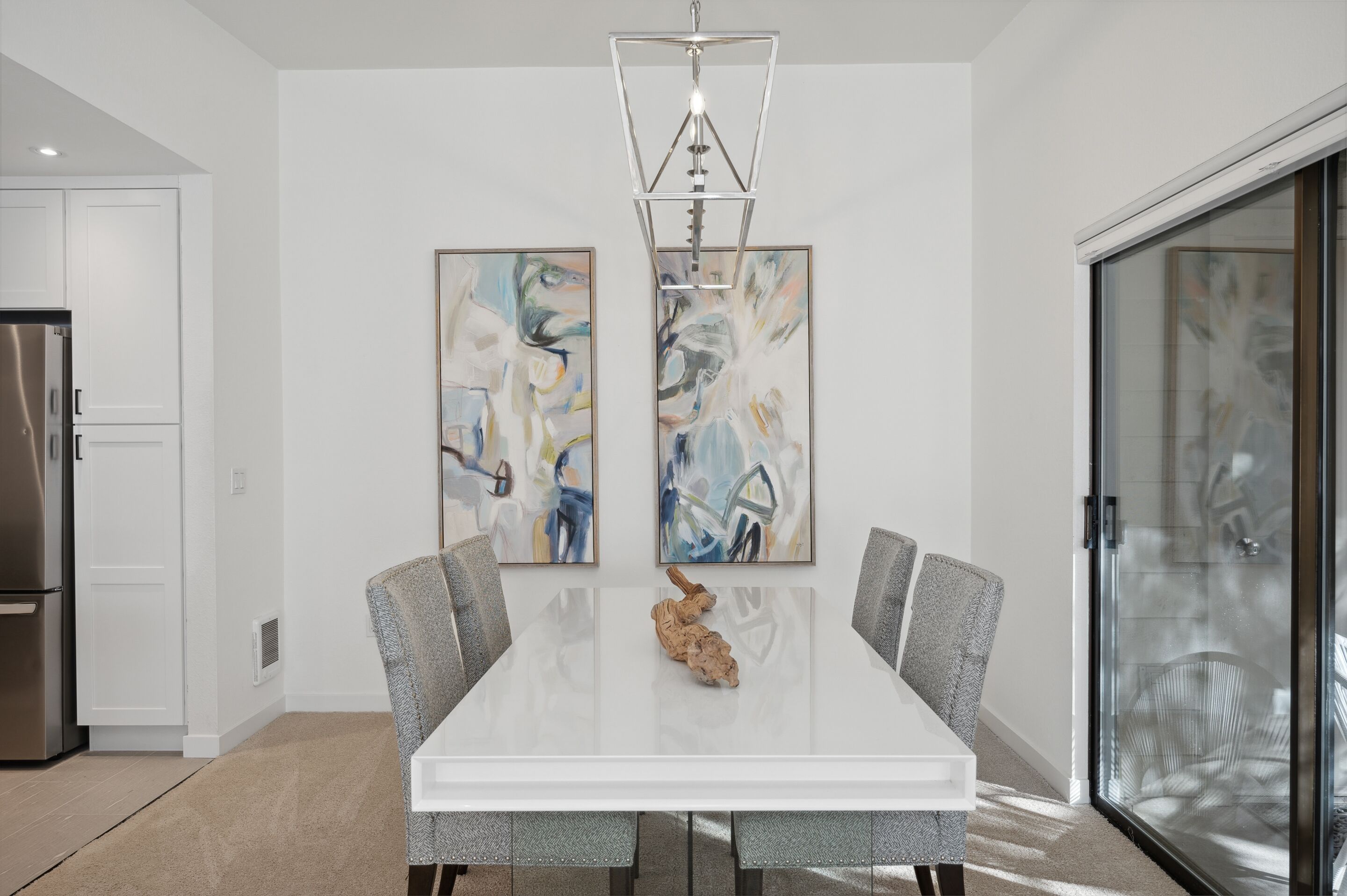 A modern dining room with a white table, grey upholstered chairs, abstract art on the walls, and a stainless steel refrigerator in the background.