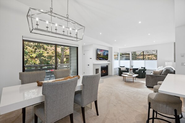 A modern living room with a fireplace, a dining area with a table and chairs, and a large window with a view of trees. - NEW Lux Bellevue 2BD 2BA Condo - Free Parking (Bellevue)