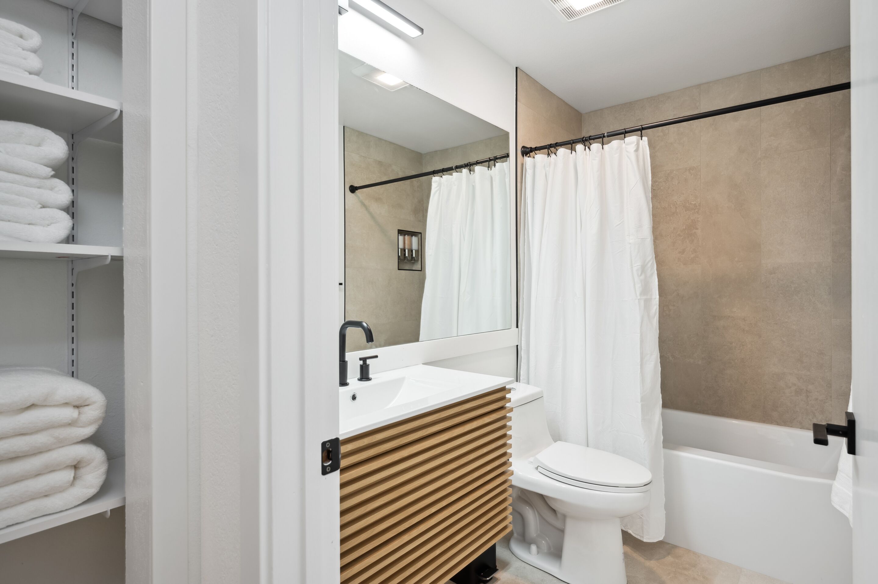 A modern bathroom with a white sink, toilet, and bathtub. There are shelves with folded towels and a mirror above the sink.