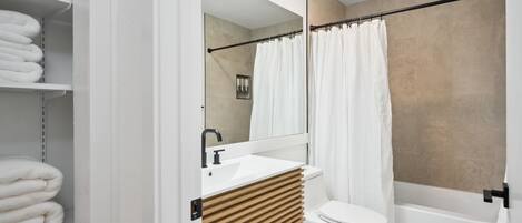 A modern bathroom with a white sink, toilet, and bathtub. There are shelves with folded towels and a mirror above the sink.