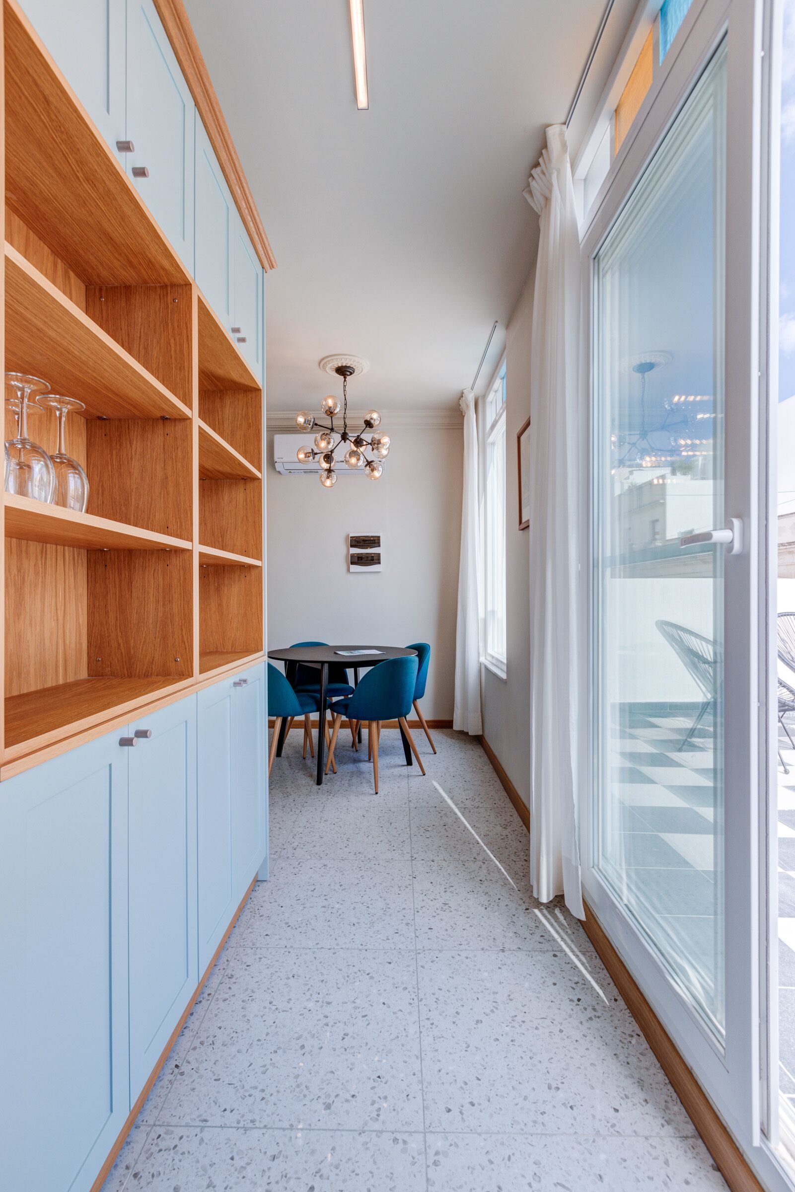 Une cuisine moderne avec des meubles de couleur bleu clair, un coin repas avec une table ronde et des chaises bleues, et une grande fenêtre offrant une vue sur la ville.