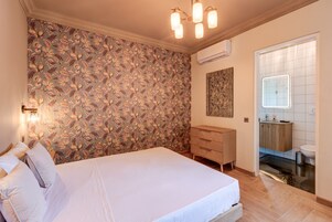 Un dormitorio con papel tapiz floral, una cama con almohadas, un tocador, un espejo y un equipo de aire acondicionado.