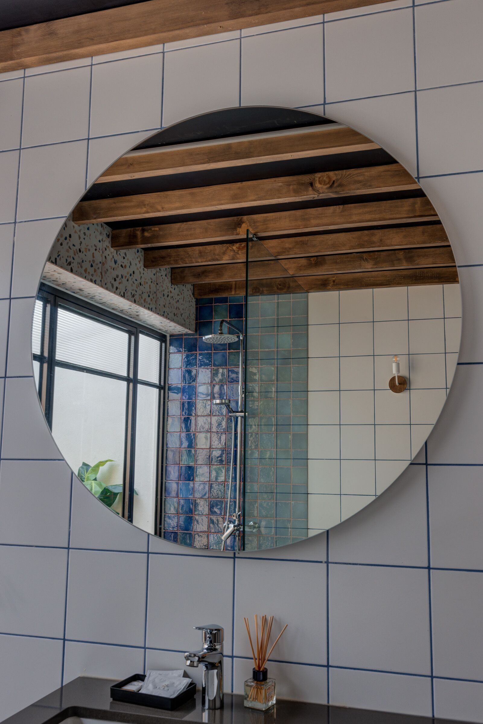 A bathroom with a large round mirror, tiled walls, a wooden ceiling, and a window.