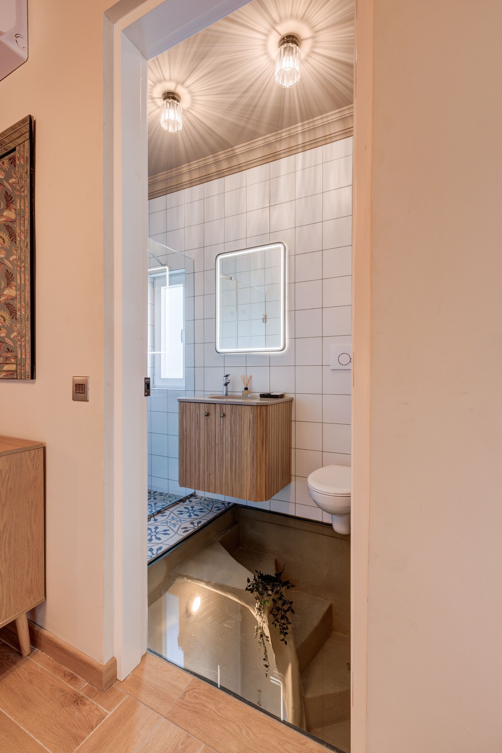 A modern bathroom with a wooden vanity, a white sink, and a mirror above it. There is a shower area with a glass door and a potted plant.