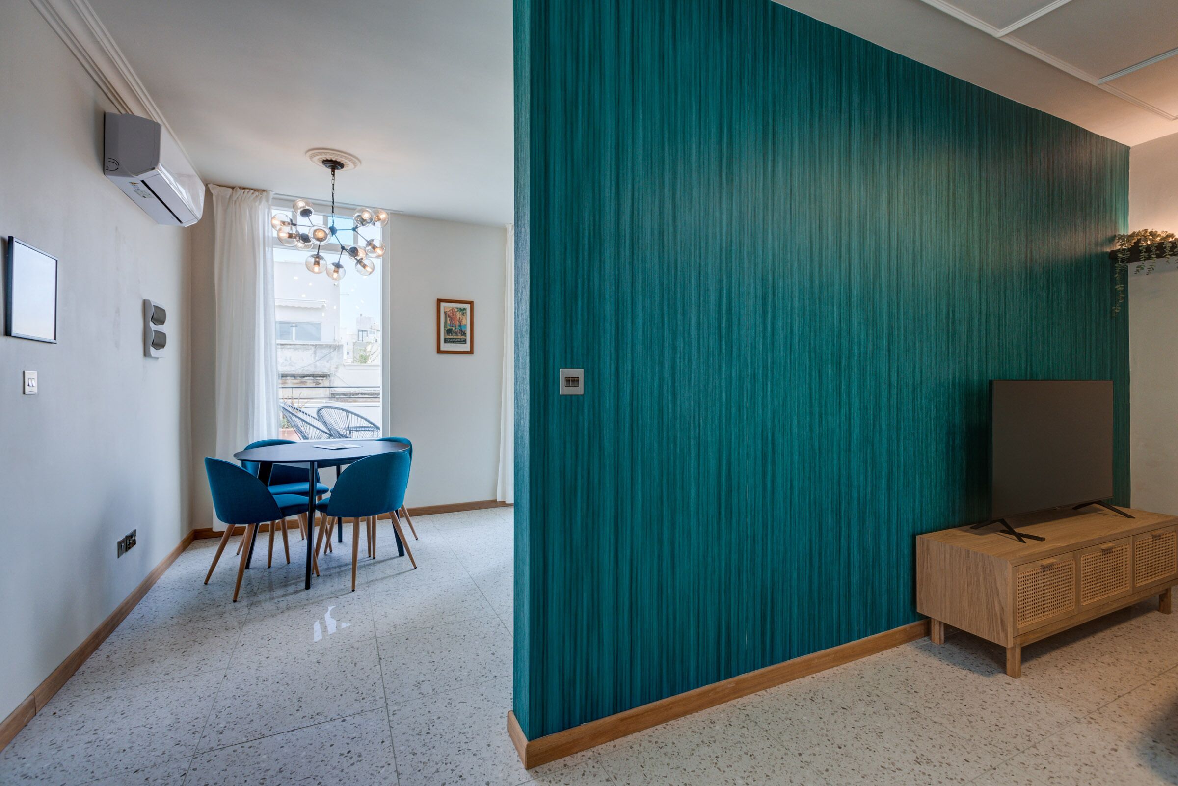 Una sala de estar moderna con papel tapiz verde azulado, una zona de comedor con sillas azules, un mueble de madera y un televisor de pantalla plana.