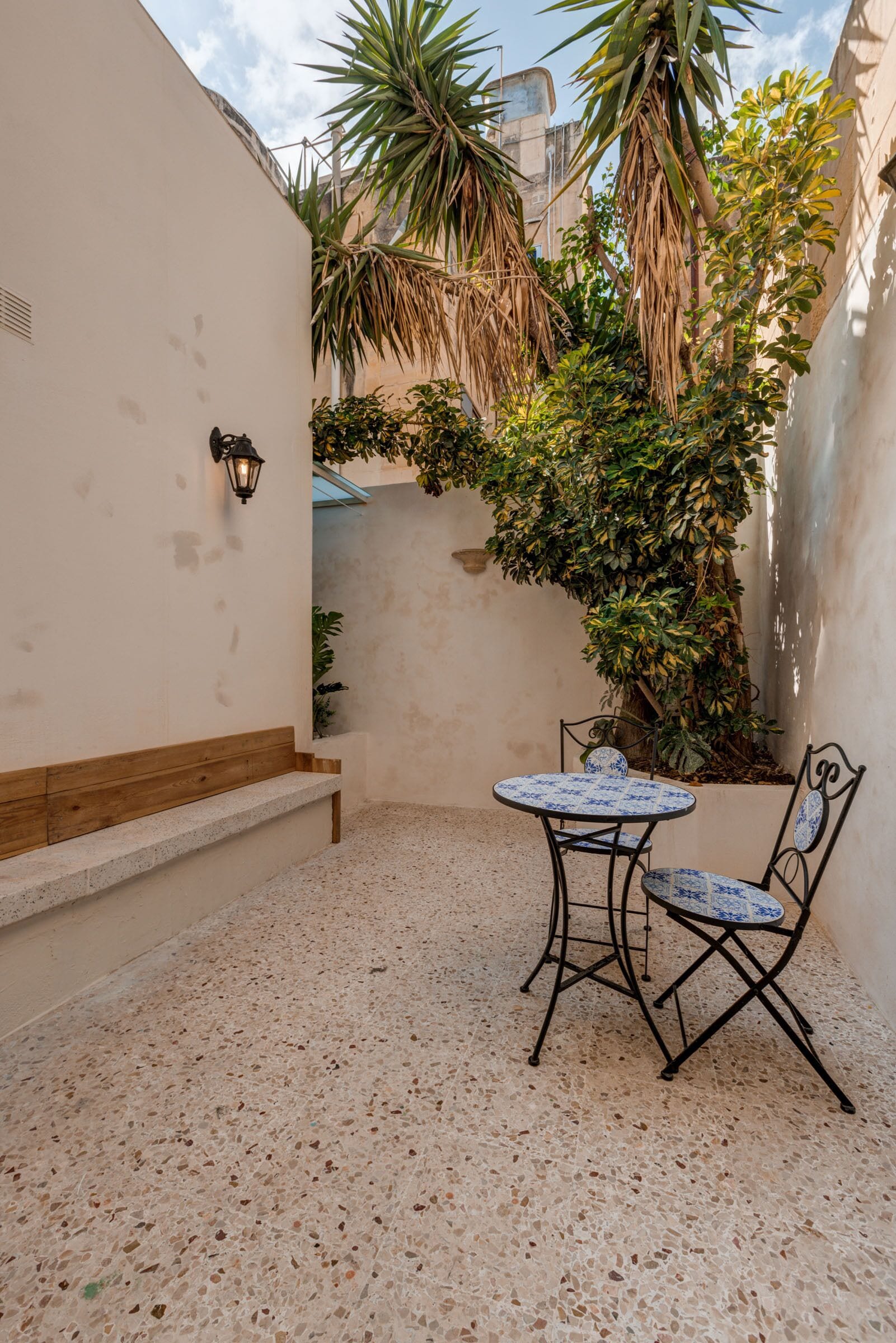 A small patio with a round table and two chairs, surrounded by plants and a white wall.