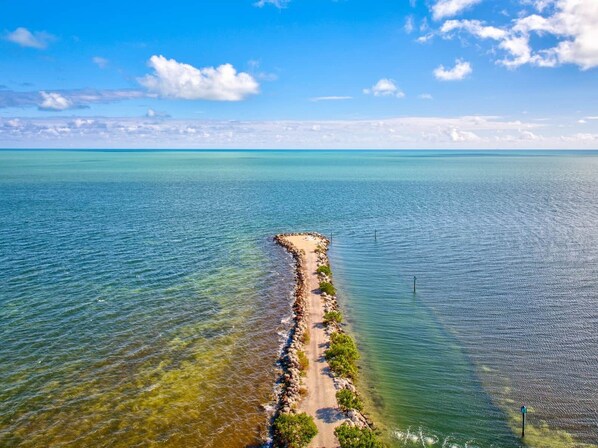 Luftaufnahme einer Küstenlandschaft mit einer schmalen Landzunge, die ins Meer führt, umgeben von klarem Wasser und blauem Himmel.