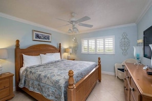Une chambre avec un lit en bois, un ventilateur de plafond, une fenêtre avec des stores, une chaise et une commode.