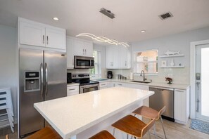 A modern kitchen with white cabinetry, stainless steel appliances, a central island with bar stools, and a window with blinds.