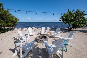 Eine Strandlandschaft mit weißen Adirondack-Stühlen, die um eine Feuerstelle angeordnet sind, mit Blick auf den Ozean.