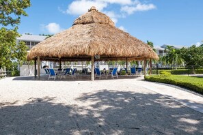 A thatched-roof pavilion with blue chairs and a gravel floor.