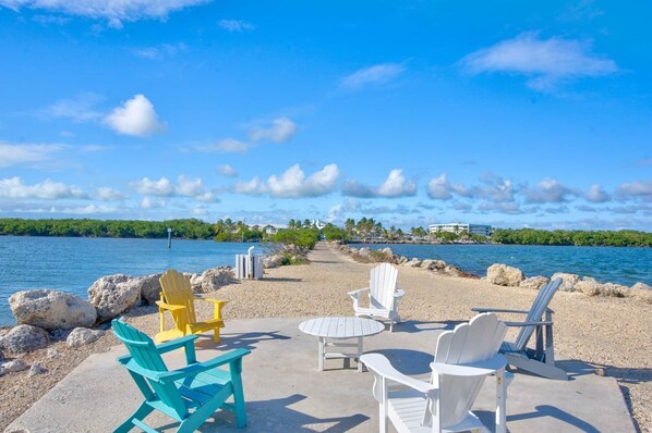 A beachside seating area with chairs and a table, overlooking a calm body of water and a distant shoreline with buildings.