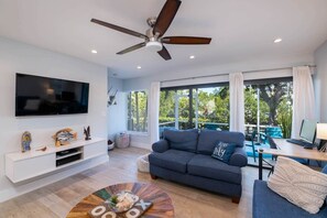 A modern living room with a flat-screen TV, a ceiling fan, a sofa, a dining area, and large windows.