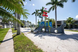 A playground with slides and a pirate ship play structure, surrounded by palm trees and a building with balconies.