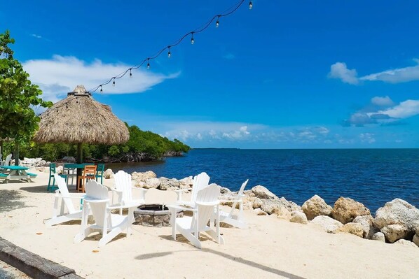 A beachside seating area with white plastic chairs and a thatched-roof table, overlooking the ocean.