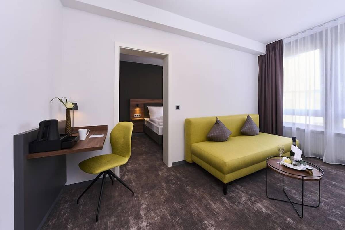 A hotel room with a green sofa, a small table with a bottle and glasses, a desk with a chair, a bed, and a nightstand with a lamp.
