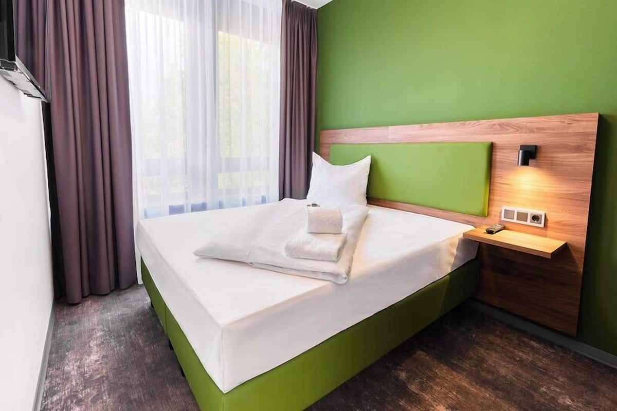 A neatly made bed in a room with a green wall, a wooden headboard, and a bedside table with a lamp.