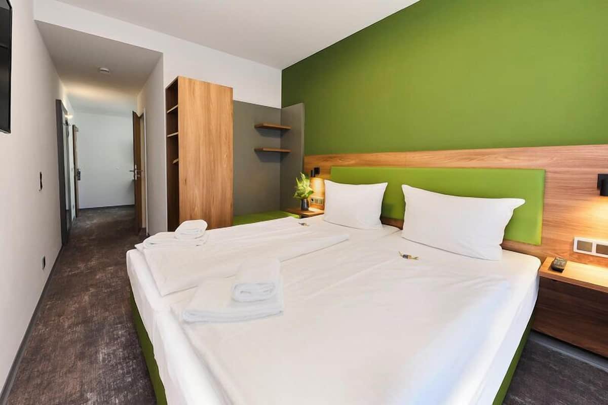 A hotel room with a large bed, a wooden wardrobe, a green accent wall, and a bedside table with a lamp.