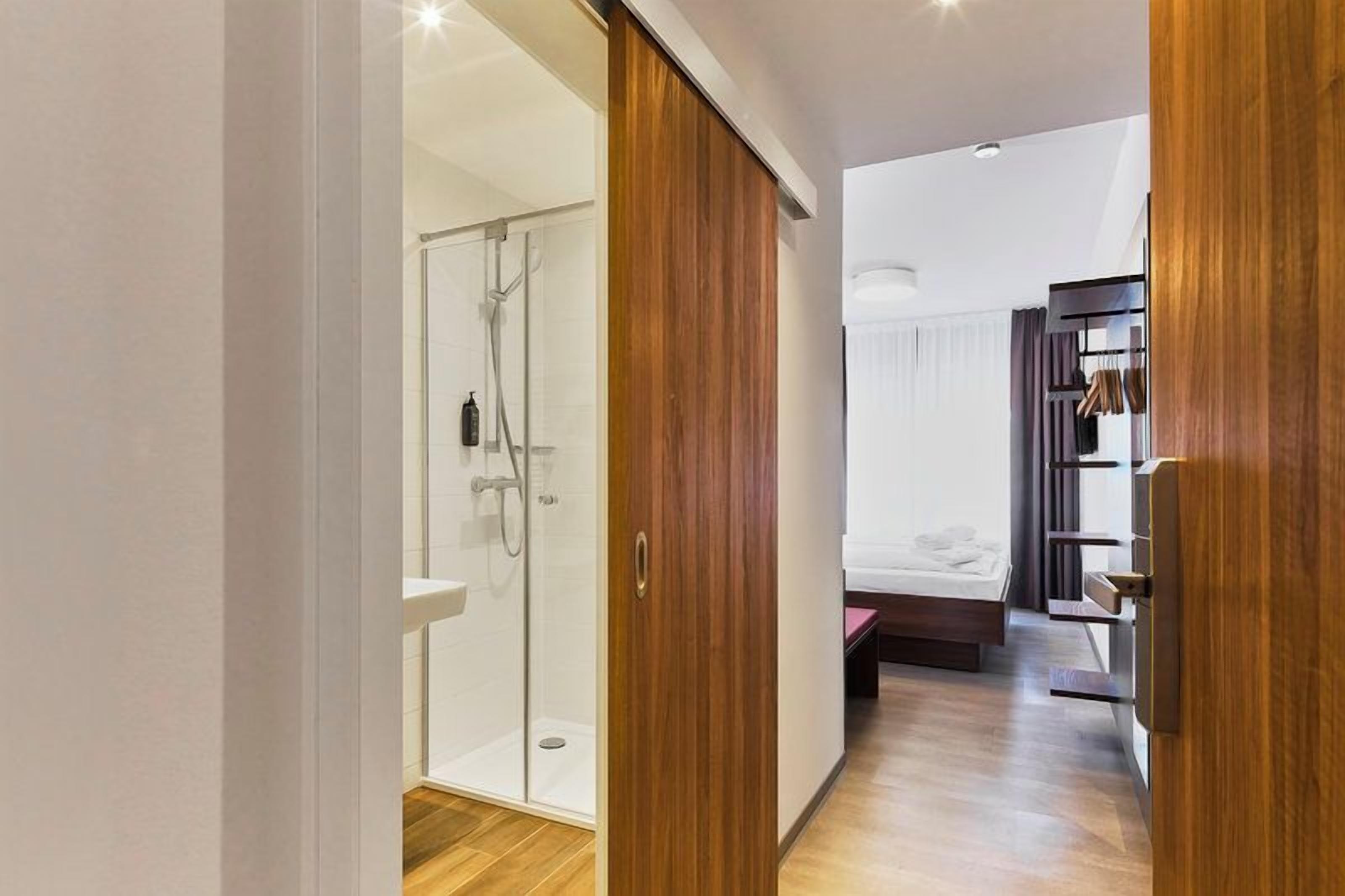 A modern bathroom with a glass shower enclosure, a wooden wardrobe, and a bed in the adjacent room.