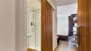 A modern bathroom with a glass shower enclosure, a wooden wardrobe, and a bed in the adjacent room.