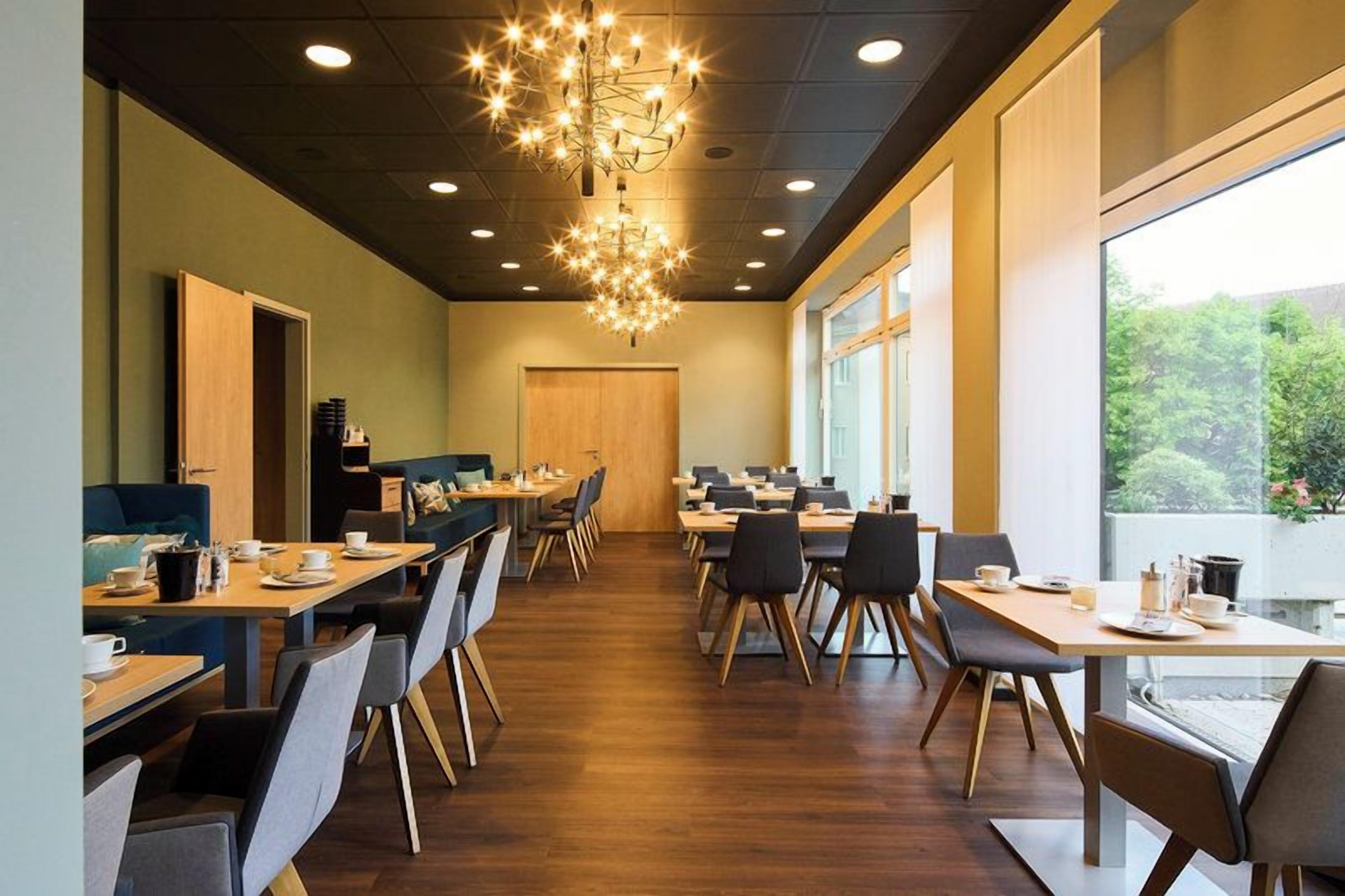 A modern dining area with wooden tables and chairs, a chandelier, and large windows offering a view of greenery.