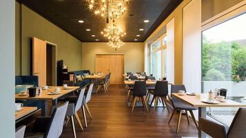 A modern dining area with wooden tables and chairs, a chandelier, and large windows offering a view of greenery.