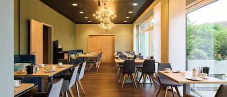 A modern dining area with wooden tables and chairs, a chandelier, and large windows offering a view of greenery.