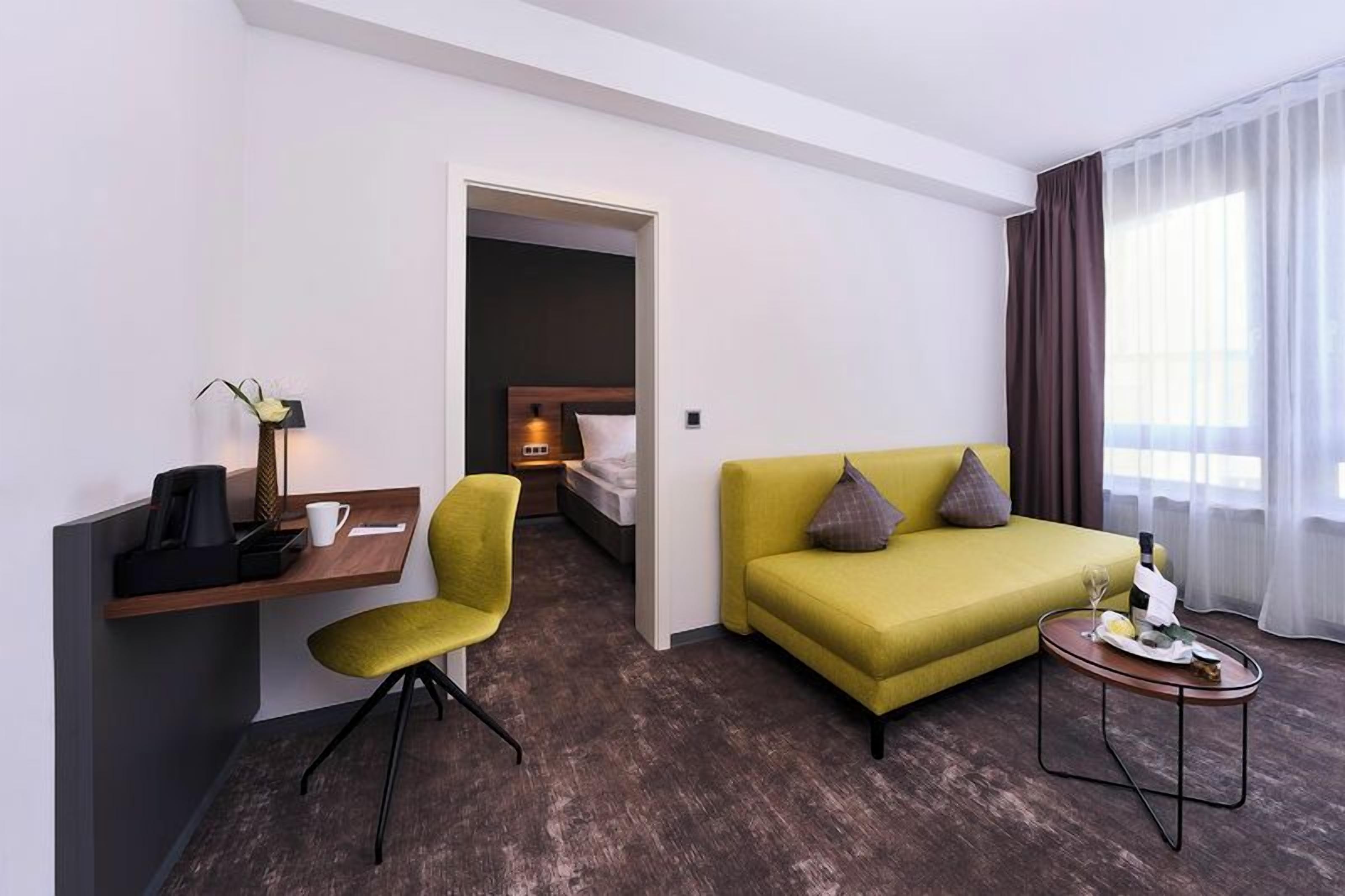 Une chambre d’hôtel avec un canapé vert, une petite table sur laquelle se trouvent une bouteille et des verres, un bureau avec une chaise, un lit et une table de chevet avec une lampe.