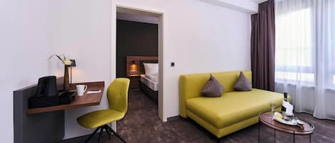 A hotel room with a desk, a yellow armchair, a sofa with cushions, a bedside table with a bottle, and a window with curtains.