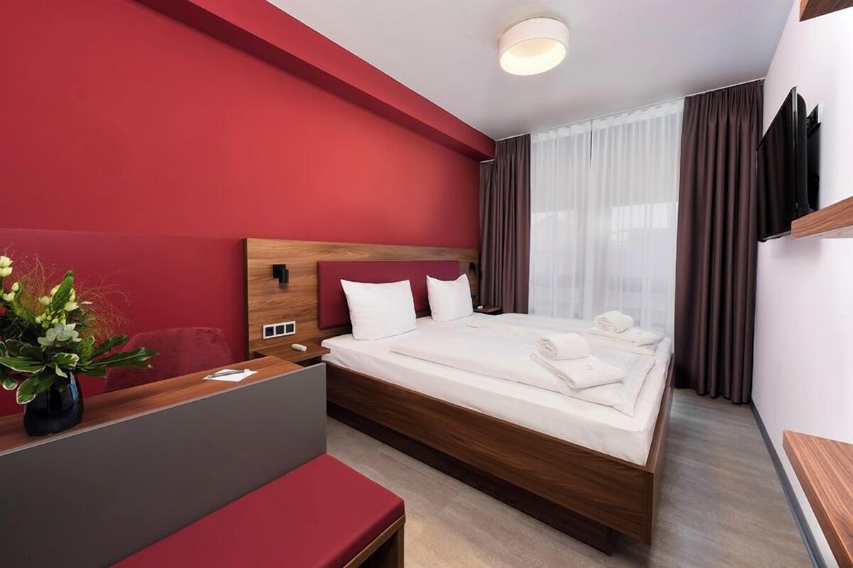 Moderni hotellihuone, jossa on suuri sänky, puinen pääty, työpöytä maljakko täynnä kukkia ja punainen tehosteseinä.