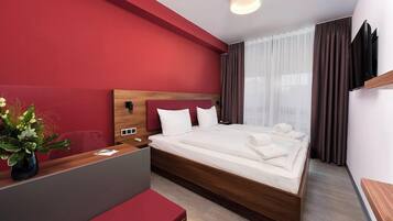 A modern hotel room with a large bed, a wooden headboard, a desk with a vase of flowers, and a red accent wall.