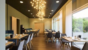 A modern dining area with wooden tables and chairs, a chandelier, and large windows offering a view of greenery. - Top Hotel Rhoenkitz Bad Kissingen (Bad Kissingen)