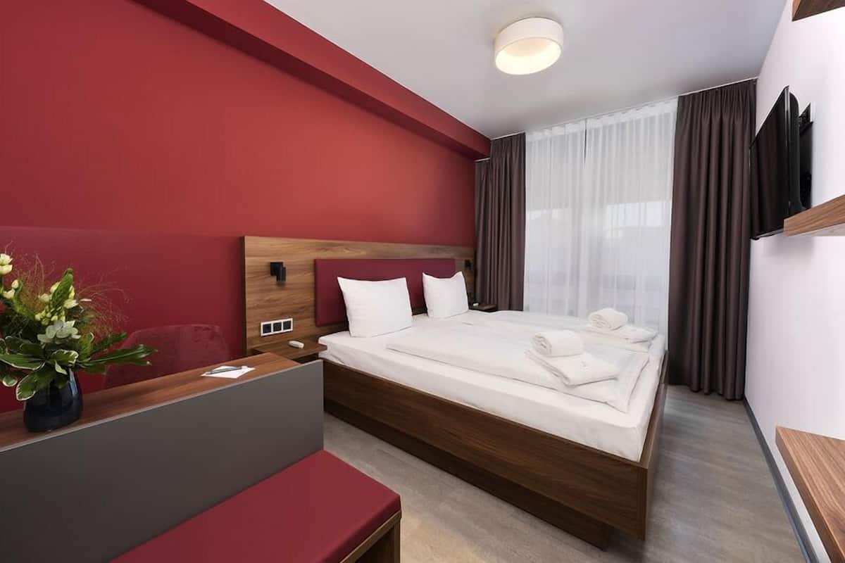 A modern hotel room with a large bed, a wooden headboard, a desk with a vase of flowers, and a red accent wall.