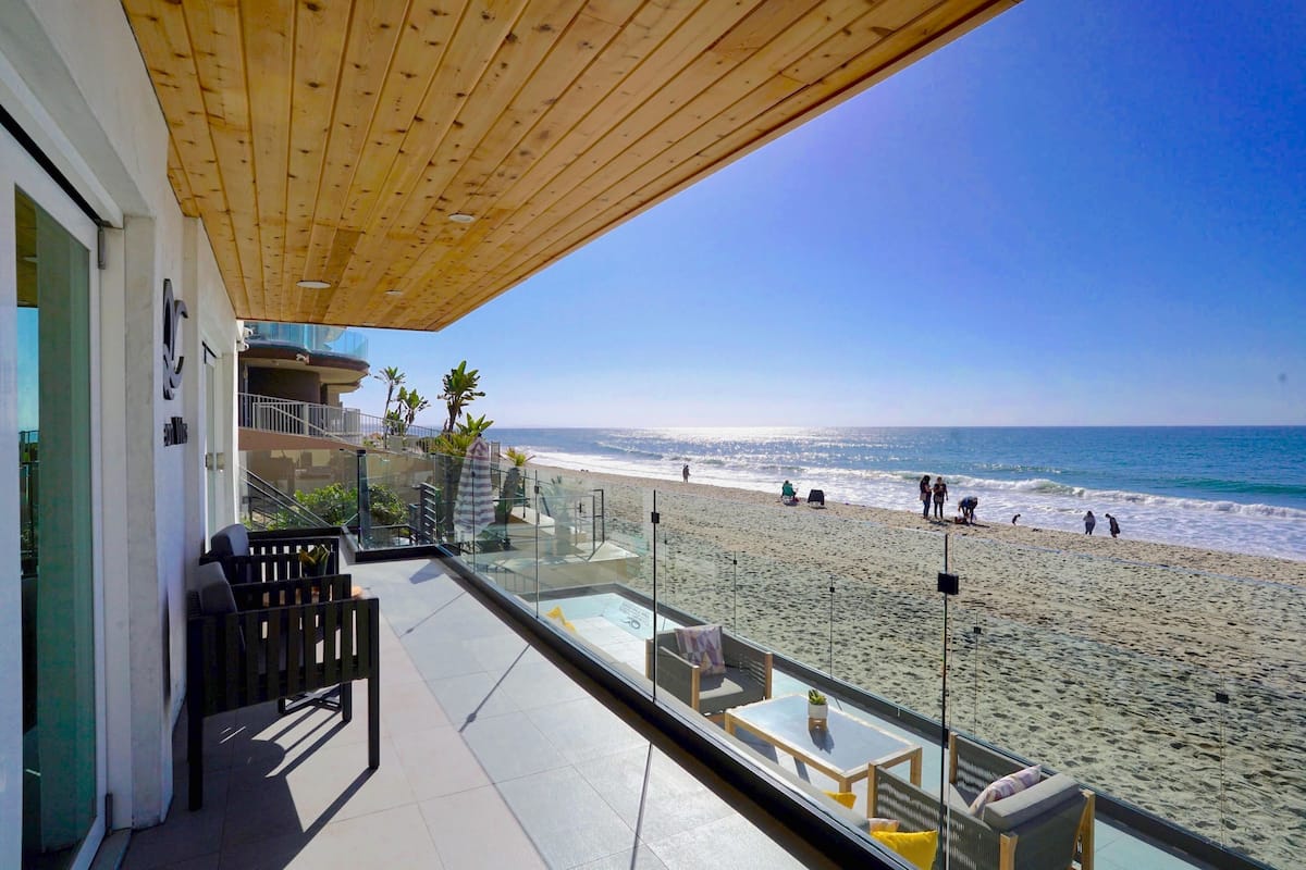 A balcony with a view of the beach, outdoor seating, and a wooden ceiling.